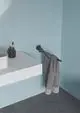 Handtuchstange in Schwarz, montiert an einer blauen Wand, hält ein graues Handtuch. Im Hintergrund sind ein Waschbecken und ein Behälter für Zahnbürsten sichtbar. Ideal Standard.